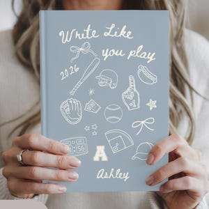 May include: A light blue journal with the text "Write Like You Play" and baseball-themed illustrations. The journal includes the name "Ashley" and the year "2026". The cover features a baseball bat, glove, and other sports-related graphics.