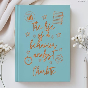 以下が含まれることがあります： オレンジ色のスクリプトで「The life of a behavior analyst Charlotte」と書かれた、ターコイズ色のノート。ノートには、星、タイマー、パズルのピースなどのオレンジ色のグラフィックがあります。Alora Studioのロゴは右上にあります。