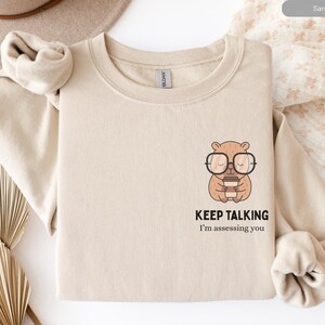 Puede incluir: Sudadera color arena con cuello redondo. Presenta un oso de dibujos animados con gafas y una taza de café. Debajo del oso está el texto "KEEP TALKING" y "I'm assessing you" en fuente negra.