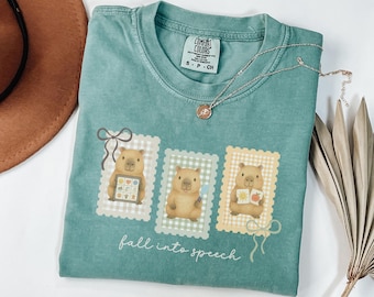 May include: A teal Comfort Colors t-shirt featuring three cartoon capybaras on postage stamp designs. Each capybara holds an item, including a tablet and an apple. The text "fall into speech" is printed at the bottom.
