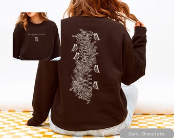 Pode incluir: Camisola de cor chocolate escuro com um design floral e borboleta branco nas costas. A frente da camisola tem o texto "the alignment artist" com um pequeno gráfico de borboleta.