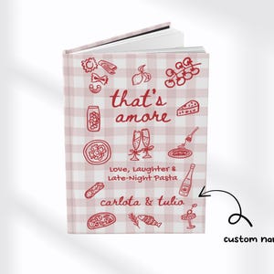 Puede incluir: Un cuaderno de tapa dura con un patr&oacute;n de cuadros rosas y blancos. La portada presenta la frase "that's amore" con ilustraciones de pasta, champ&aacute;n y otros alimentos italianos. Tambi&eacute;n se incluye el texto "Love, Laughter & Late-Night Pasta".