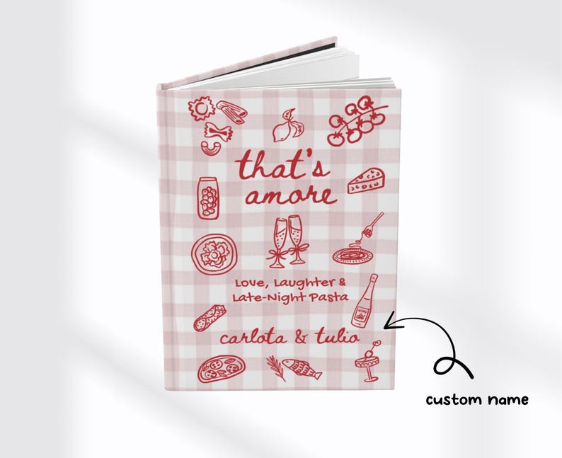 Puede incluir: Un cuaderno de tapa dura con un patr&oacute;n de cuadros rosas y blancos. La portada presenta la frase "that's amore" con ilustraciones de pasta, champ&aacute;n y otros alimentos italianos. Tambi&eacute;n se incluye el texto "Love, Laughter & Late-Night Pasta".