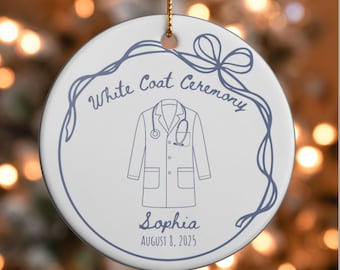 Könnte beinhalten: Weißer Keramikschmuck mit blauem Umriss eines Arztmantels und Stethoskops. Der Text "White Coat Ceremony", "Sophia" und "August 8, 2025" ist ebenfalls blau. Ein goldener Faden ist zum Aufhängen befestigt.