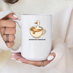 May include: White ceramic mug featuring a cartoon duck in a coffee cup design. The duck is white with an orange beak and feet. The mug has the words "preferred reinforcer" printed in black.