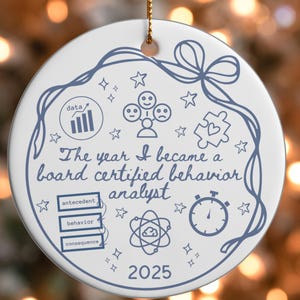 Op de afbeelding: Ronde witte keramische kerstversiering met een blauw ontwerp. De tekst luidt "The year I became a board certified behavior analyst" en het jaar 2025. Inclusief gerelateerde iconen en woorden.