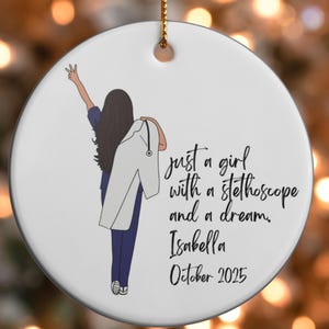 May include: A white ceramic Christmas ornament with a gold hanging loop. It depicts a person in scrubs with a lab coat and stethoscope. The text reads "just a girl with a stethoscope and a dream. Isabella October 2025."