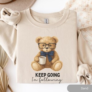 Puede incluir: Sudadera color arena con una ilustración en acuarela de un oso de peluche con gafas y pajarita, sosteniendo una taza de café. El texto "KEEP GOING I'm following" está impreso debajo.