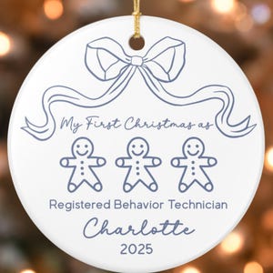 May include: White ceramic Christmas ornament with a gold string for hanging. The ornament features a blue bow and ribbon design at the top, with the text "My First Christmas as" above three gingerbread people. Below the gingerbread people is the text "Registered Behavior Technician Charlotte 2025."