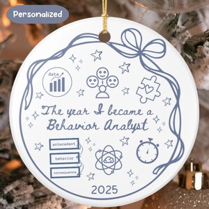 Op de afbeelding: Witte keramische ornament met een blauw ontwerp en gouden touw. Het ornament heeft de tekst "The year I became a Behavior Analyst" en het jaar 2025. Het bevat verschillende iconen en het woord "Personalized".