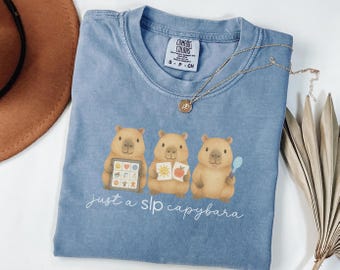 May include: A light blue Comfort Colors t-shirt featuring three cartoon capybaras. Each capybara holds an item, including a communication board, a book, and a spoon. The text "just a slp capybara" is printed below the capybaras.