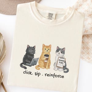 Puede incluir: Camiseta de color crema con un gráfico de tres gatos de dibujos animados. Un gato lleva gafas y usa un portátil, otro sostiene una taza de café y el tercero sostiene un letrero que dice "Behavior Plan". El texto "click. sip. reinforce" está debajo.