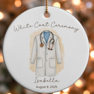May include: White ceramic ornament with a watercolor illustration of a doctor's coat and stethoscope. The text "White Coat Ceremony" is at the top, with the name "Isabella" and the date "August 8, 2025" below the coat.