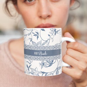 May include: White ceramic mug with a floral design in blue and gray. The mug features the text "RBT Nicole" on a gray band. The floral pattern includes leaves and flowers, creating an elegant and personalized look. The mug has a white handle.
