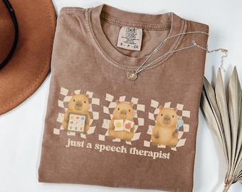 May include: Brown Comfort Colors t-shirt with the text "just a speech therapist" and three cartoon capybaras. Each capybara holds a different item: a tablet, a book, and a bubble wand. The background features a checkered pattern.
