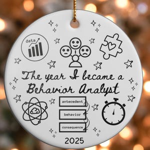 Op de afbeelding: Ronde witte keramische kerstversiering met zwarte illustraties en tekst. Het ontwerp bevat een datadiagram, gezichten, een puzzelstukje, sterren en een stopwatch. De tekst luidt "The year I became a Behavior Analyst" en "2025". Bevat "antecedent", "behavior" en "consequence".