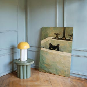 May include: A yellow and white table lamp with a mushroom-shaped top sits on a green side table. A painting of a black cat in a bathtub is propped against the wall. The room has light blue walls and a wooden floor.