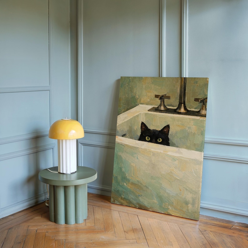 May include: A yellow and white table lamp with a mushroom-shaped top sits on a green side table. A painting of a black cat in a bathtub is propped against the wall. The room has light blue walls and a wooden floor.
