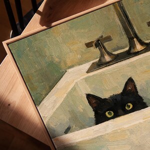 May include: Oil painting depicting a black cat peering over a white sink's edge. The artwork features a light green and beige colour scheme, with visible brushstrokes. The painting is framed in a light-coloured wooden frame.