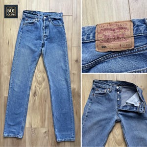 Op de afbeelding: Lichtblauwe denim jeans met een knoopsluiting. Het Levi Strauss & Co.-label is zichtbaar. De jeans is maat 26x34. Het 501 Club-logo staat linksboven.