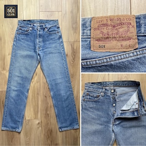 Könnte beinhalten: Hellblaue Jeans mit Knopfleiste. Die Jeans haben ein klassisches Fünf-Taschen-Design und einen braunen Lederaufnäher mit dem Text "LEVI STRAUSS & CO." und "501". Die Jeans werden auf einer Holzoberfläche präsentiert.