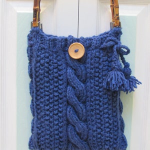 May include: A blue knitted tote bag with a wooden handle and a wooden button. The bag has a cable knit pattern and tassels.