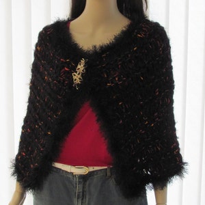 May include: A black fuzzy shawl with a red shirt underneath. The shawl has a decorative gold brooch.