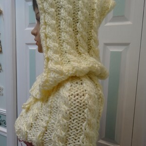 May include: A cream-colored knitted hooded cape with a cable knit pattern. The cape is made of soft yarn and has a loose fit.