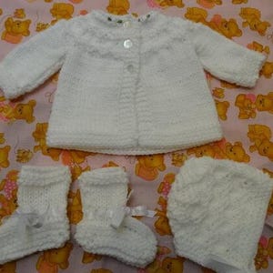 May include: A white knitted baby outfit set including a cardigan, hat, and booties. The set is made of soft, white yarn and features a simple, classic design.