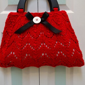 May include: A red crocheted handbag with a black ribbon and a white button. The bag has a black wooden handle.