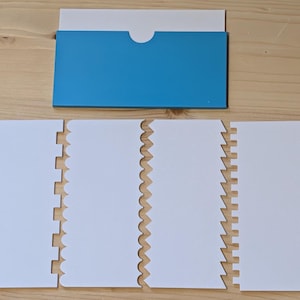 May include: A set of white cake scrapers with various edge designs, including straight, zigzag, and scalloped patterns. A blue and white envelope is positioned above the scrapers on a wooden surface. These tools are used for cake decorating.