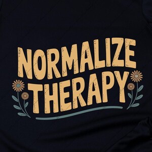 May include: Black t-shirt with the words "NORMALIZE THERAPY" in a light brown, stylized font. The text is decorated with small flowers and leaves. The shirt is on a light wood surface with sunglasses, a bracelet, and jeans.