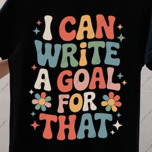 Könnte beinhalten: Schwarzes T-Shirt mit dem Text "I CAN WRITE A GOAL FOR THAT" in einer farbenfrohen Retro-Schriftart. Das Design beinhaltet Blumen und Sterne. Das T-Shirt wird von einer Person präsentiert.