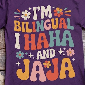 May include: A purple t-shirt with the text "I'M BILINGUAL I HAHA AND JAJA" in colorful, retro-style lettering. The design includes flowers and stars. The shirt is displayed on a wooden surface with autumn leaves and boots.