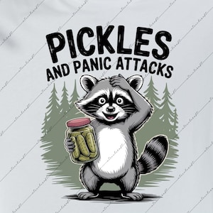 May include: White t-shirt featuring the text "Pickles and Panic Attacks" and a cartoon raccoon holding a jar of pickles. The raccoon is grey, black, and white, with a red lid on the jar. The background shows a forest of green trees.