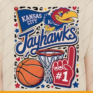Puede incluir: Camiseta color crema con un gráfico de baloncesto. El diseño incluye una pelota de baloncesto, un aro y un dedo de espuma rojo con el número uno. También está presente el texto "Kansas City Jayhawks".