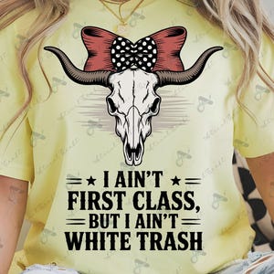 May include: A yellow t-shirt with a cow skull graphic, long horns, and a red bow with black polka dots. The text reads "I AIN'T FIRST CLASS, BUT I AIN'T WHITE TRASH". The shirt is worn by a person.