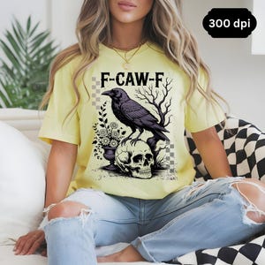 May include: A light yellow t-shirt featuring a graphic of a black crow perched on a skull, with the text "F-CAW-F". The design includes floral elements and bare tree branches. The shirt is worn with blue jeans.