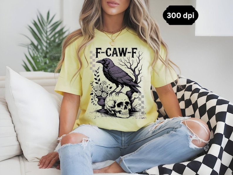 May include: A light yellow t-shirt featuring a graphic of a black crow perched on a skull, with the text "F-CAW-F". The design includes floral elements and bare tree branches. The shirt is worn with blue jeans.