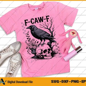 May include: Pink t-shirt with a graphic of a crow perched on a skull, with the text "F-CAW-F". The design includes a floral arrangement and a tree. Silver sneakers, a pink plush toy, and a pink tube are also in the image.