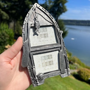 May include: A silver-toned metal picture frame shaped like a rowboat with two rectangular photo slots. The frame features molded details including two oars on the sides and a small fish charm hanging from the top. The inserts read Sixtrees.