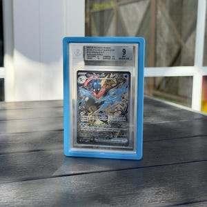 Könnte beinhalten: Eine bewertete Pokémon-Karte mit Greninja ex, in einem blauen Halter. Die Karte zeigt eine farbenfrohe Illustration von Greninja mit dem Text "Greninja ex 300". Der Halter hat eine Bewertung von 9 und enthält Details zum Zustand der Karte.
