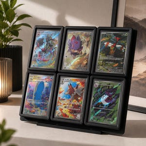 May include: A black plastic display frame holding six individual trading cards in protective cases. The cards feature colourful Japanese illustrations of various creatures and characters, arranged in a two-by-three grid format on a stand.