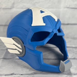 May include: A blue plastic Captain America helmet with a white 'A' on the front. The helmet has a gray chin strap and a white wing on the side.