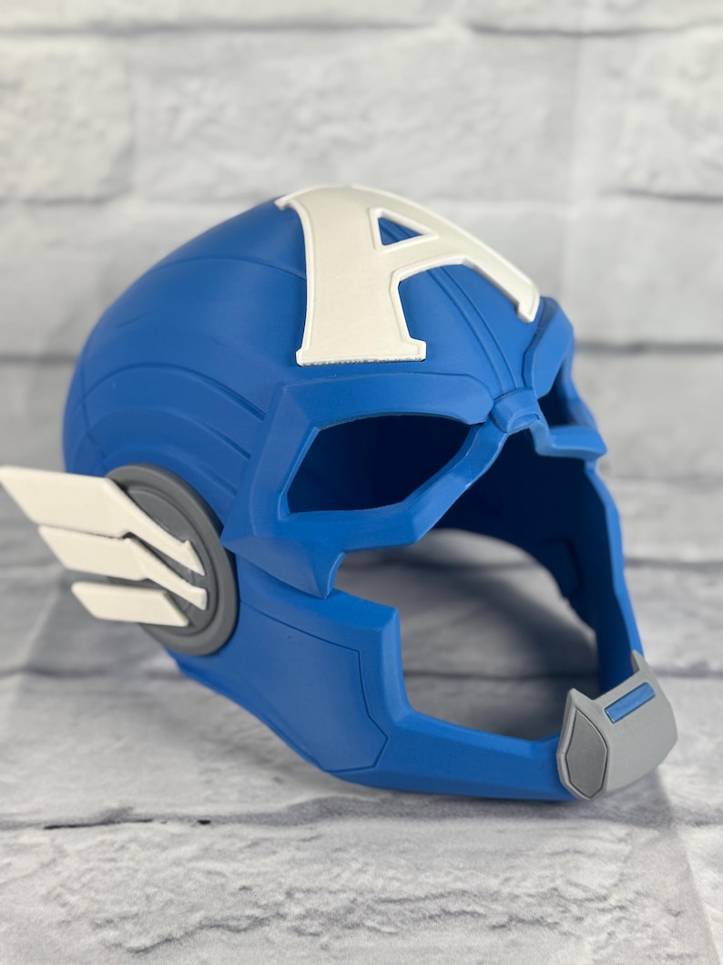 May include: A blue plastic Captain America helmet with a white 'A' on the front. The helmet has a gray chin strap and a white wing on the side.