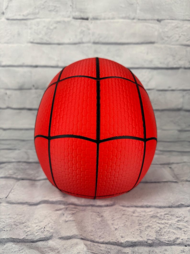 May include: A spherical red ball featuring a black web pattern across its surface. The ball has a textured finish with a repeating honeycomb-like pattern within each section created by the black lines, resembling a classic superhero costume design.