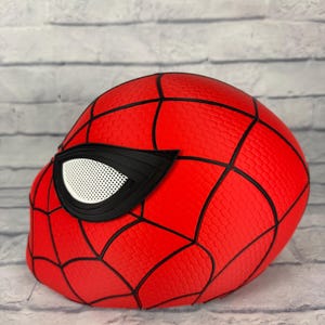 May include: A bright red full-head mask featuring a textured honeycomb pattern across the surface. The mask is accented with bold black webbing lines and a large, angular eye piece with a white mesh lens covering the eye opening.