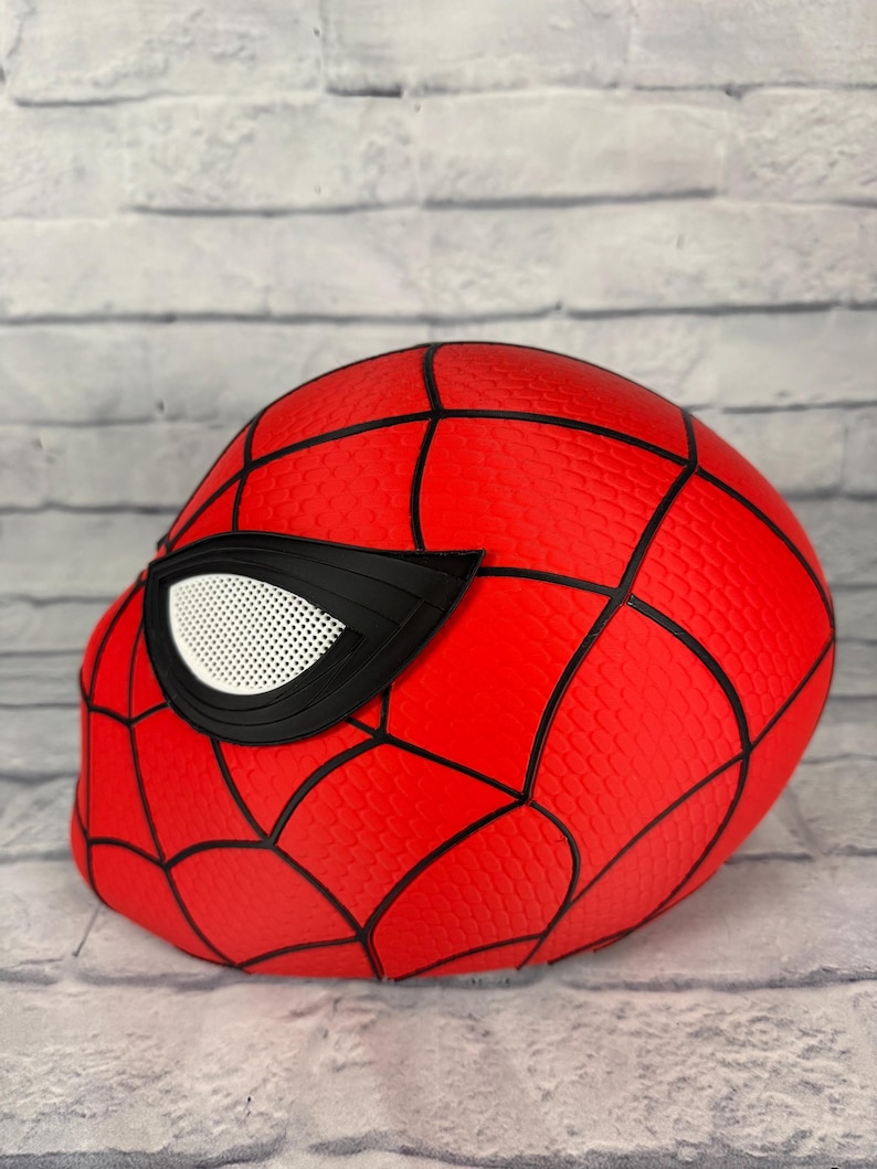 May include: A bright red full-head mask featuring a textured honeycomb pattern across the surface. The mask is accented with bold black webbing lines and a large, angular eye piece with a white mesh lens covering the eye opening.