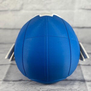 May include: Blue and white plastic football helmet with a white stripe around the top. The helmet is sitting on a white brick background.