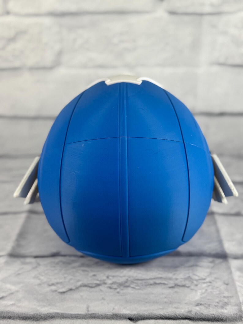 May include: Blue and white plastic football helmet with a white stripe around the top. The helmet is sitting on a white brick background.